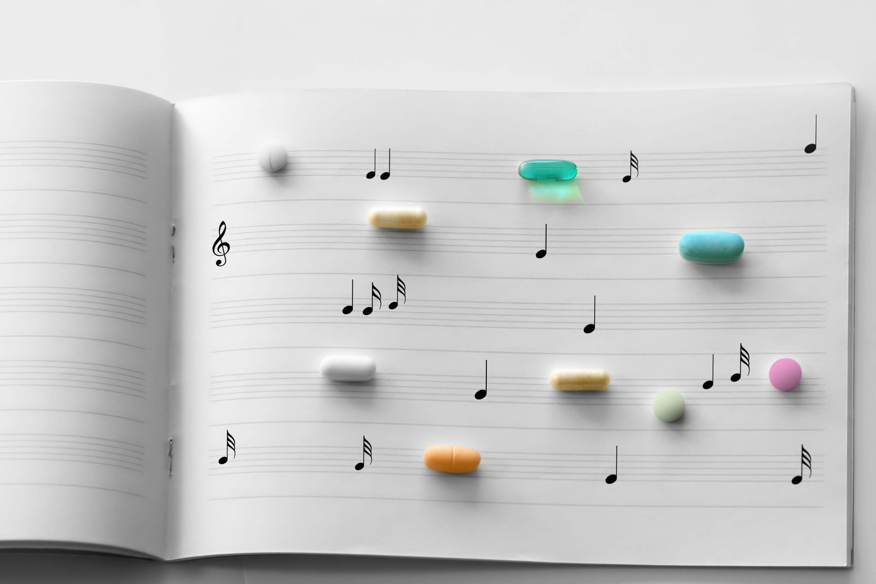 Ein aufgeschlagenes Notenheft mit Musiknoten auf den Notenlinien ist mit unterschiedlich geformten und farbigen Tabletten und Kapseln belegt. Die Medikamente sind wie einzelne Notensymbole über die Seiten verteilt und veranschaulichen die Verbindung von Musik und medizinischer Therapie, beispielsweise im Kontext von Musiktherapie oder Gesundheit.