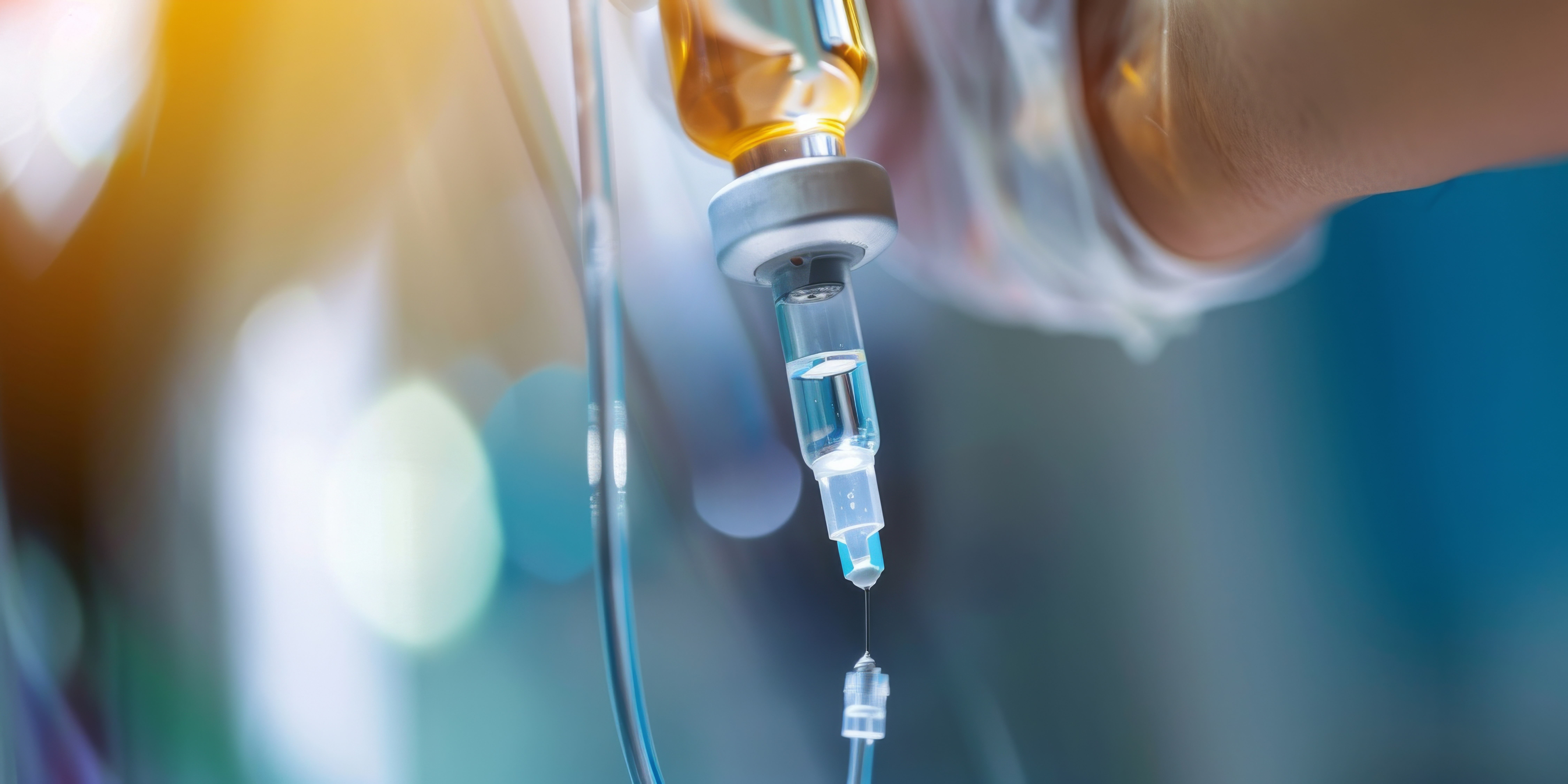 Nahaufnahme eines intravenösen Infusionssystems mit einer Glasflasche und einer Tropfkammer, durch die Flüssigkeit in einen Infusionsschlauch gelangt. Der Hintergrund ist unscharf und in kühlen Blau- sowie warmen Goldtönen gehalten. Die Darstellung steht für Infusionstherapie, Medikamentengabe und klinische Versorgung im Gesundheitswesen.