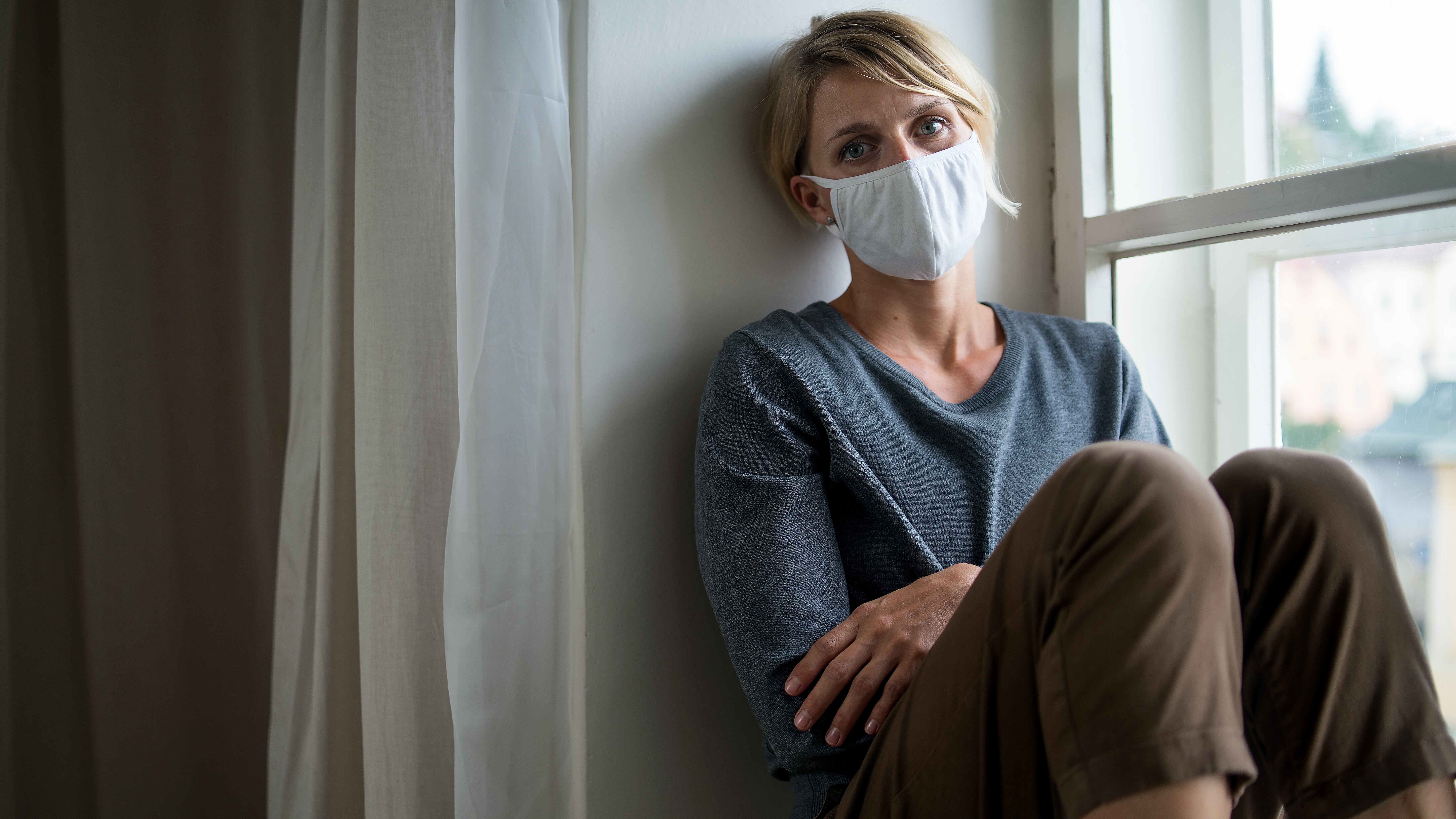 Person mit Mund-Nasen-Schutz sitzt neben einem Fenster in häuslicher Umgebung.