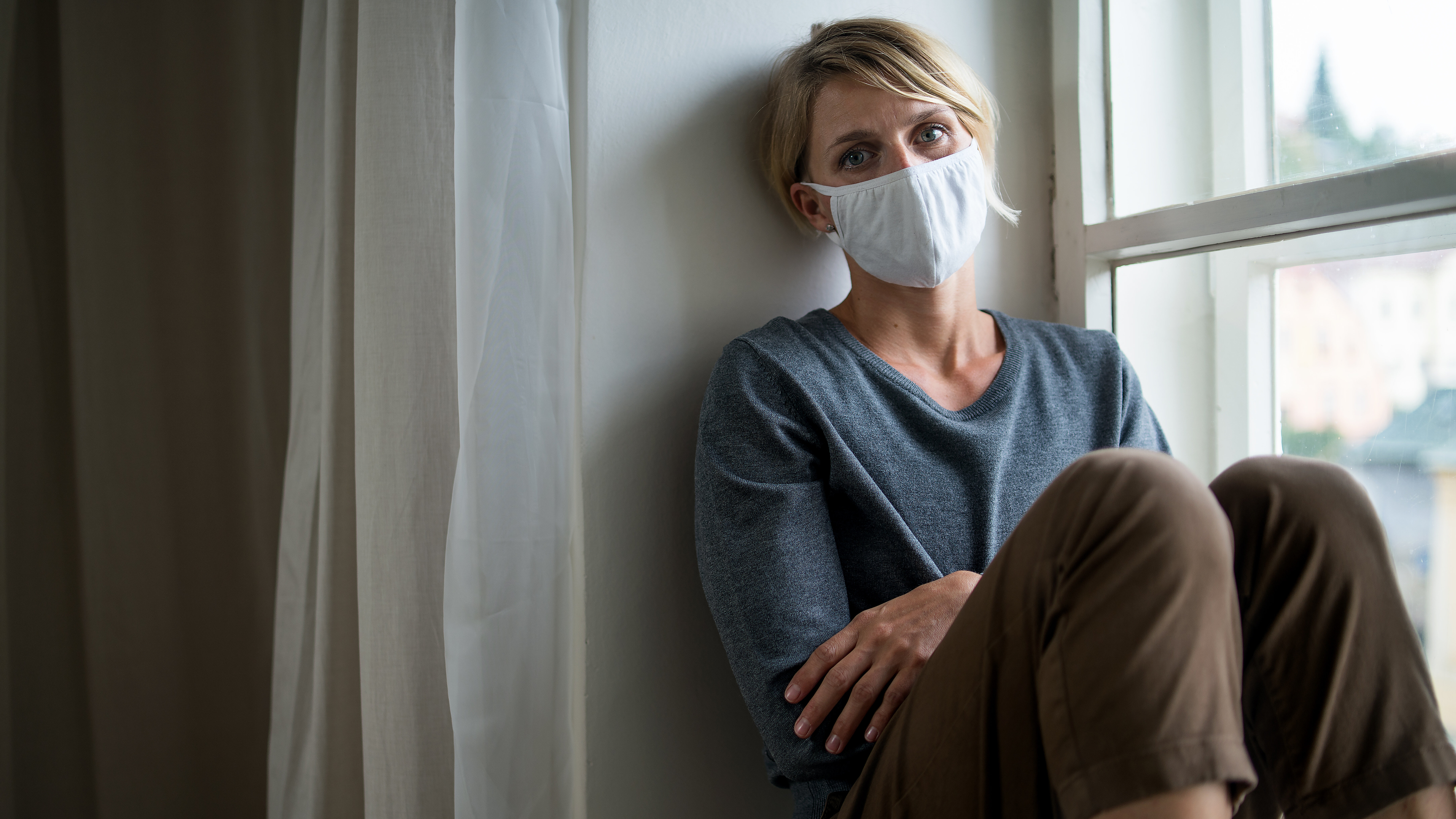 Person mit Mund-Nasen-Schutz sitzt neben einem Fenster in häuslicher Umgebung.