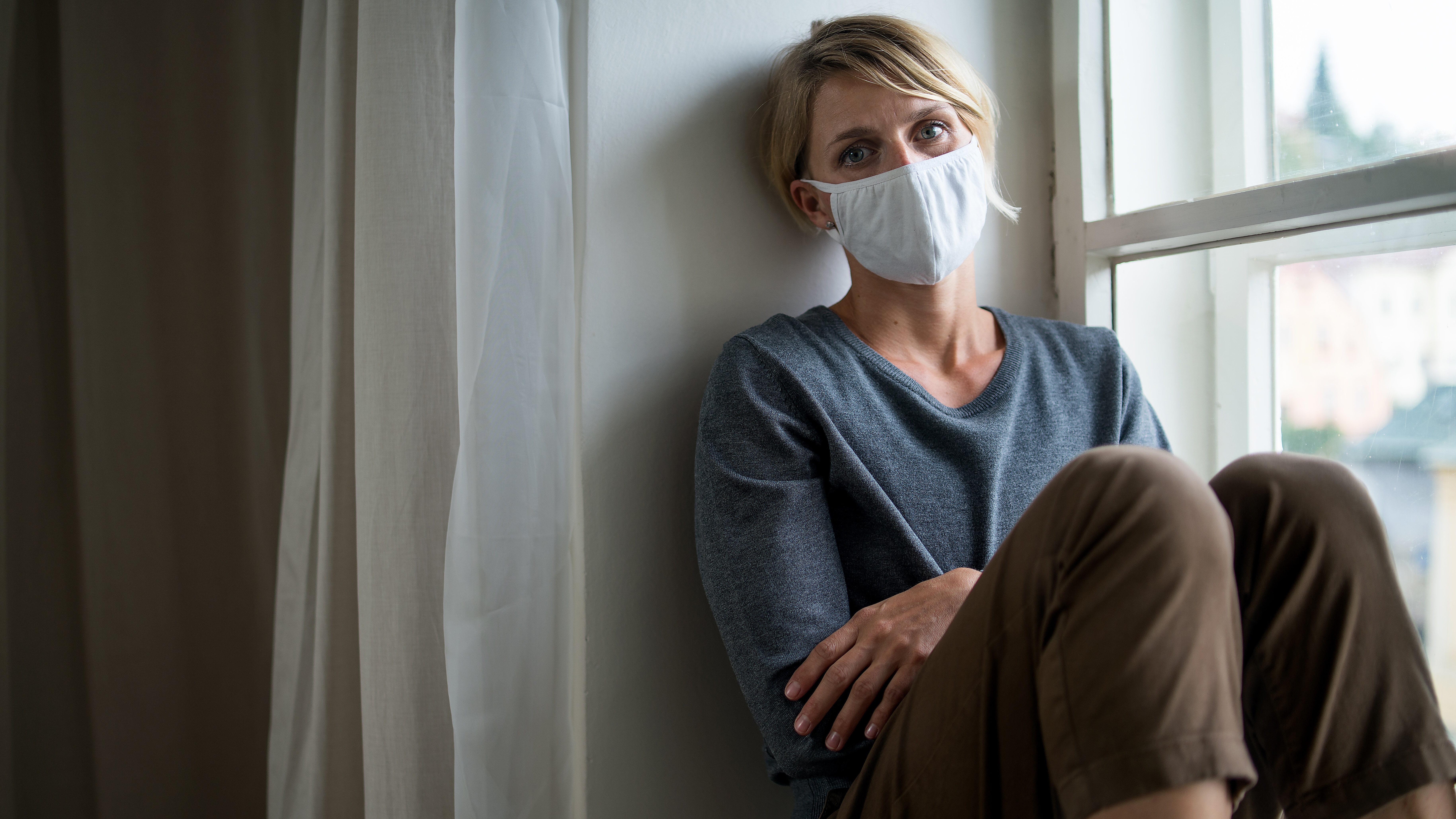 Person mit Mund-Nasen-Schutz sitzt neben einem Fenster in häuslicher Umgebung.