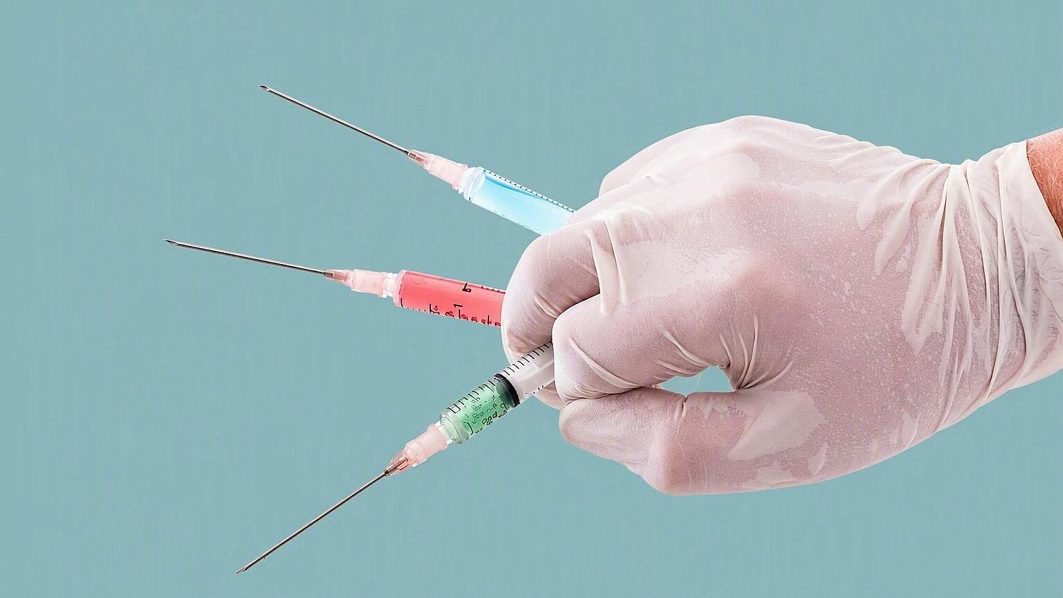 Nahaufnahme einer behandschuhten Hand, die drei Spritzen mit unterschiedlich gefärbten Flüssigkeiten hält. Die Spritzen sind fächerförmig angeordnet und vor einem hellen Hintergrund dargestellt. Das Bild symbolisiert Impfungen, Injektionen, medizinische Behandlungen sowie den Einsatz von Arzneimitteln im Gesundheitswesen.