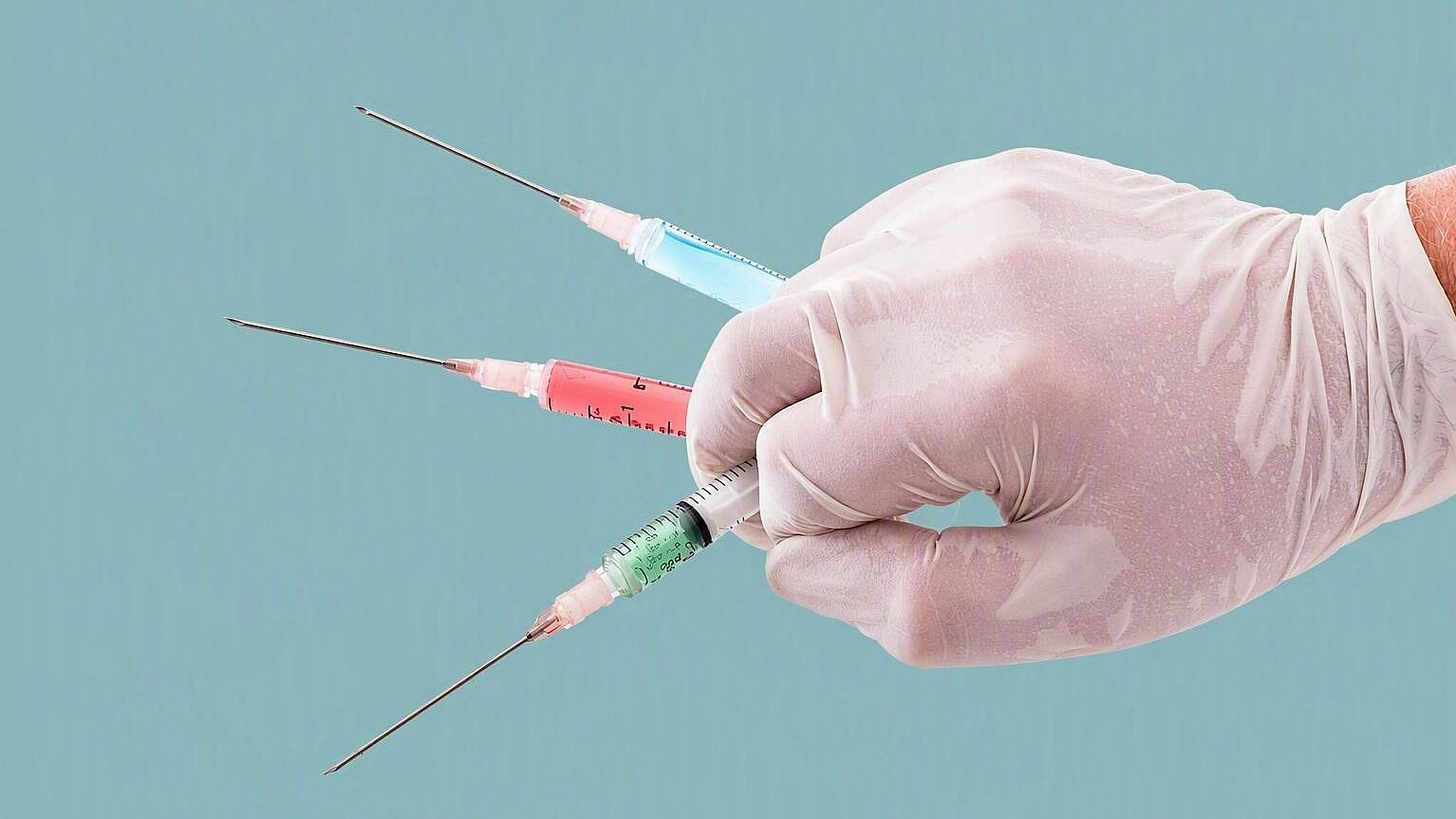 Nahaufnahme einer behandschuhten Hand, die drei Spritzen mit unterschiedlich gefärbten Flüssigkeiten hält. Die Spritzen sind fächerförmig angeordnet und vor einem hellen Hintergrund dargestellt. Das Bild symbolisiert Impfungen, Injektionen, medizinische Behandlungen sowie den Einsatz von Arzneimitteln im Gesundheitswesen.