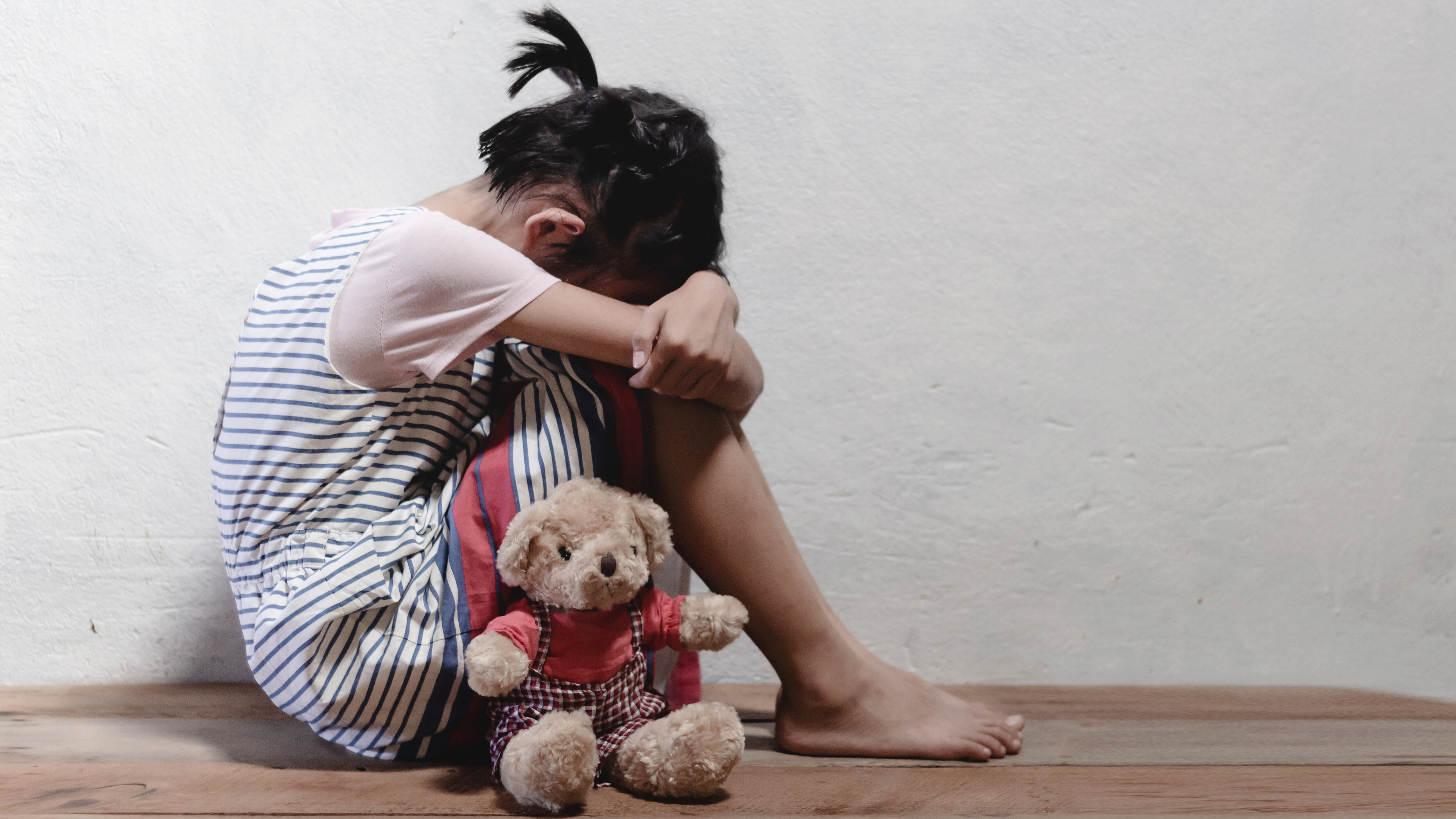 Kind sitzt mit einem Teddybären auf dem Boden und lehnt an einer Wand.