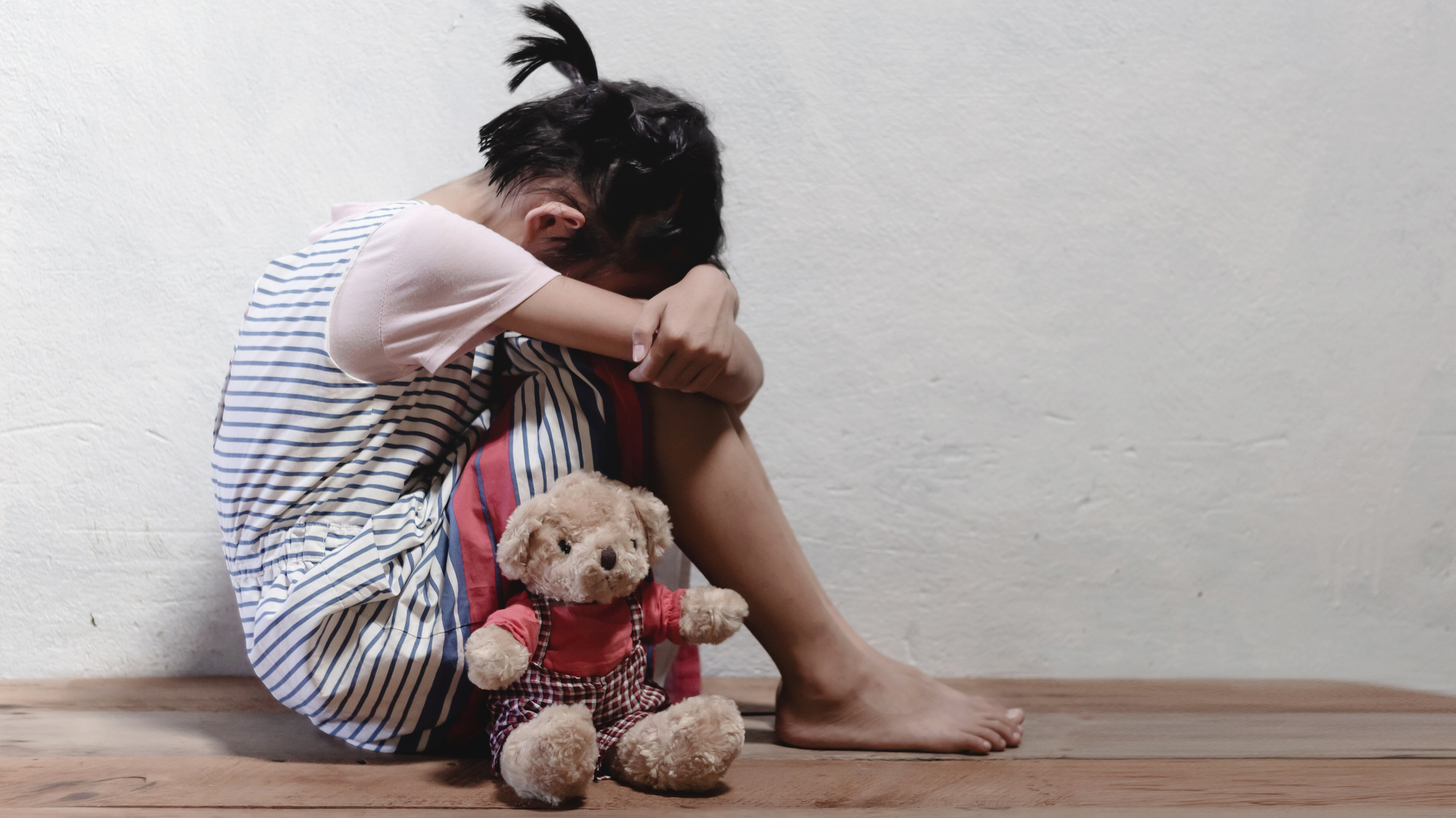 Child sitting on the floor with a teddy bear against a wall.