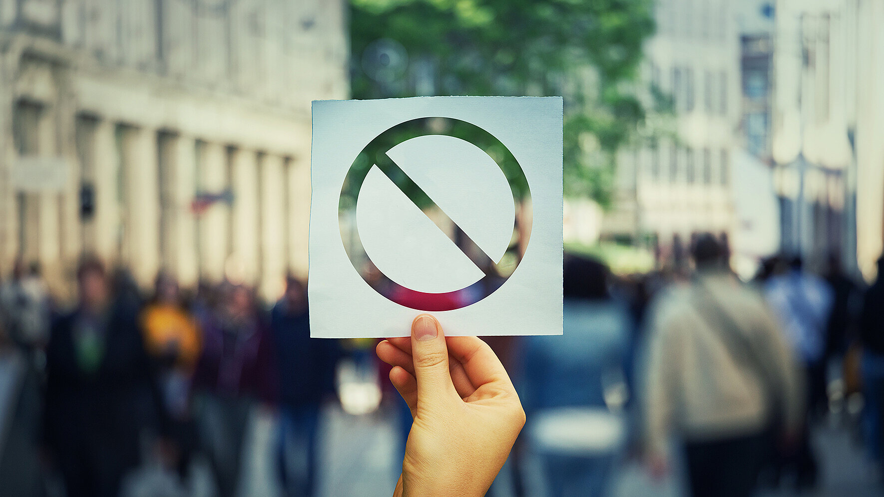 A hand holds a sign displaying a prohibition symbol in front of a blurred street scene with pedestrians. The image symbolizes restrictions, prohibitions, regulations, or the prevention of certain activities in public spaces.