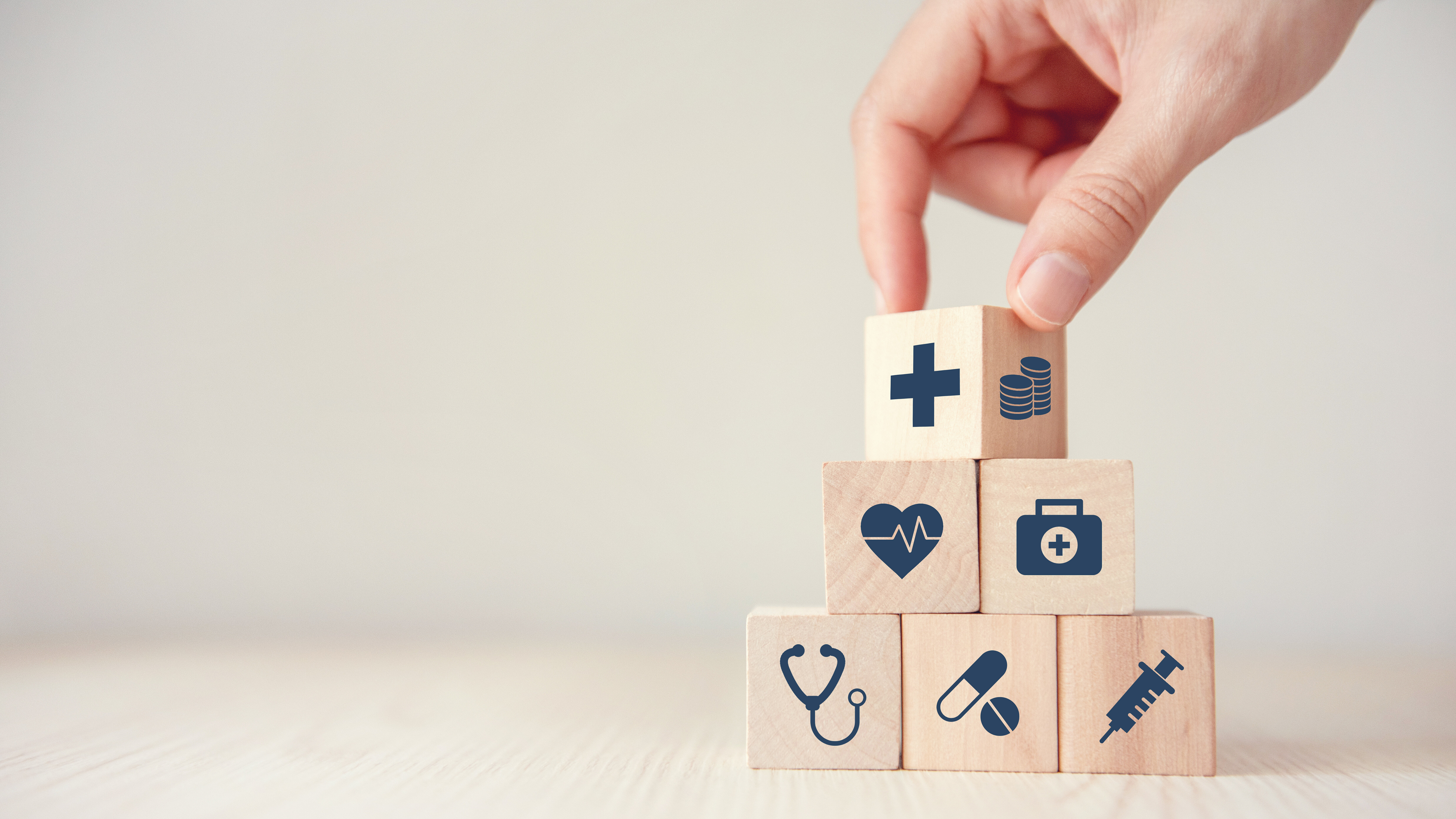 A hand stacks wooden blocks displaying healthcare-related symbols, including a stethoscope, heartbeat, medication, syringe, first-aid kit, medical cross, and coins. The image symbolizes the organisation and financing of healthcare services, as well as the different components of a healthcare system.