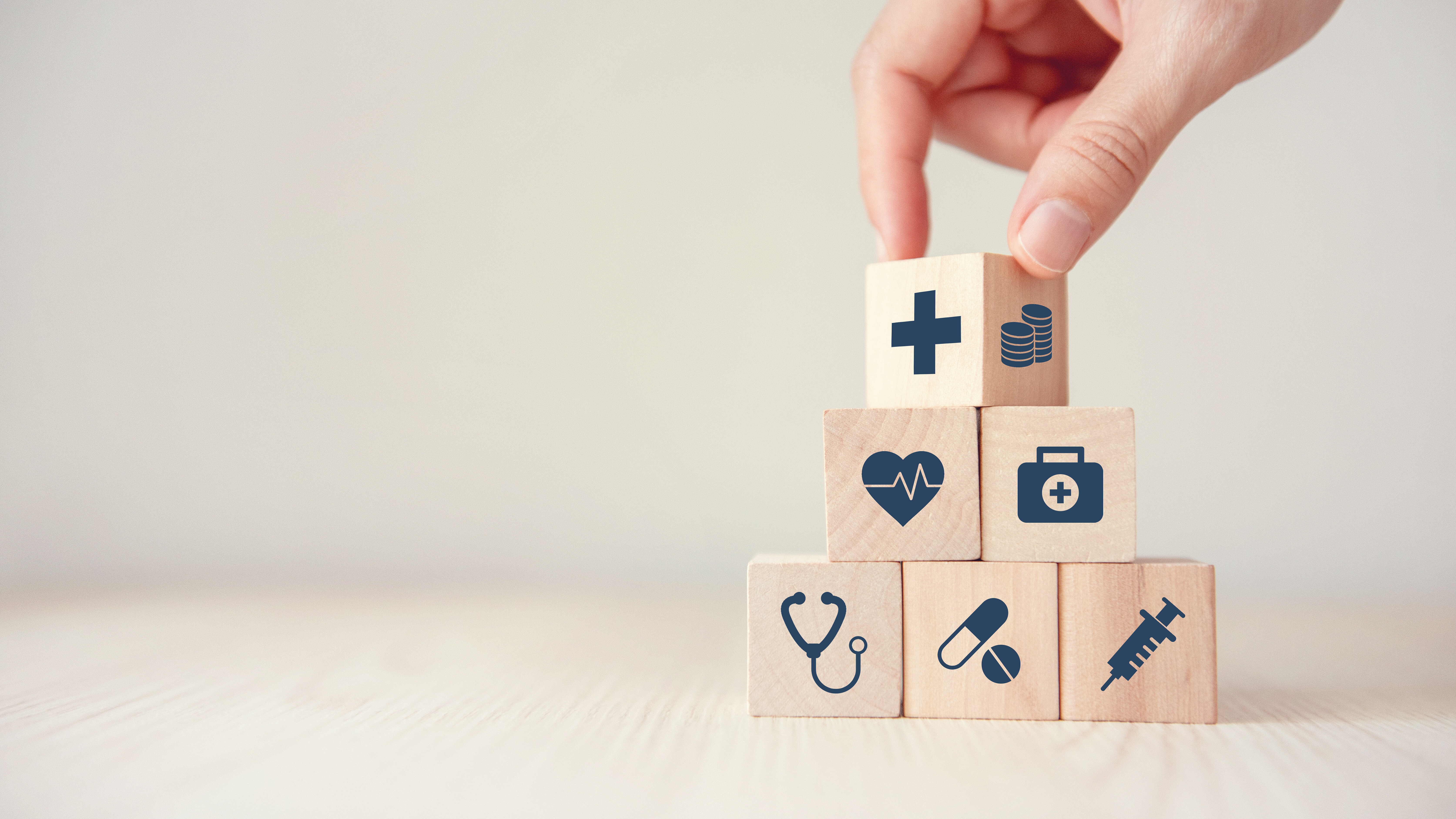A hand stacks wooden blocks with healthcare symbols, including a cross, heartbeat, medical kit, stethoscope, medication, and syringe, into a pyramid.