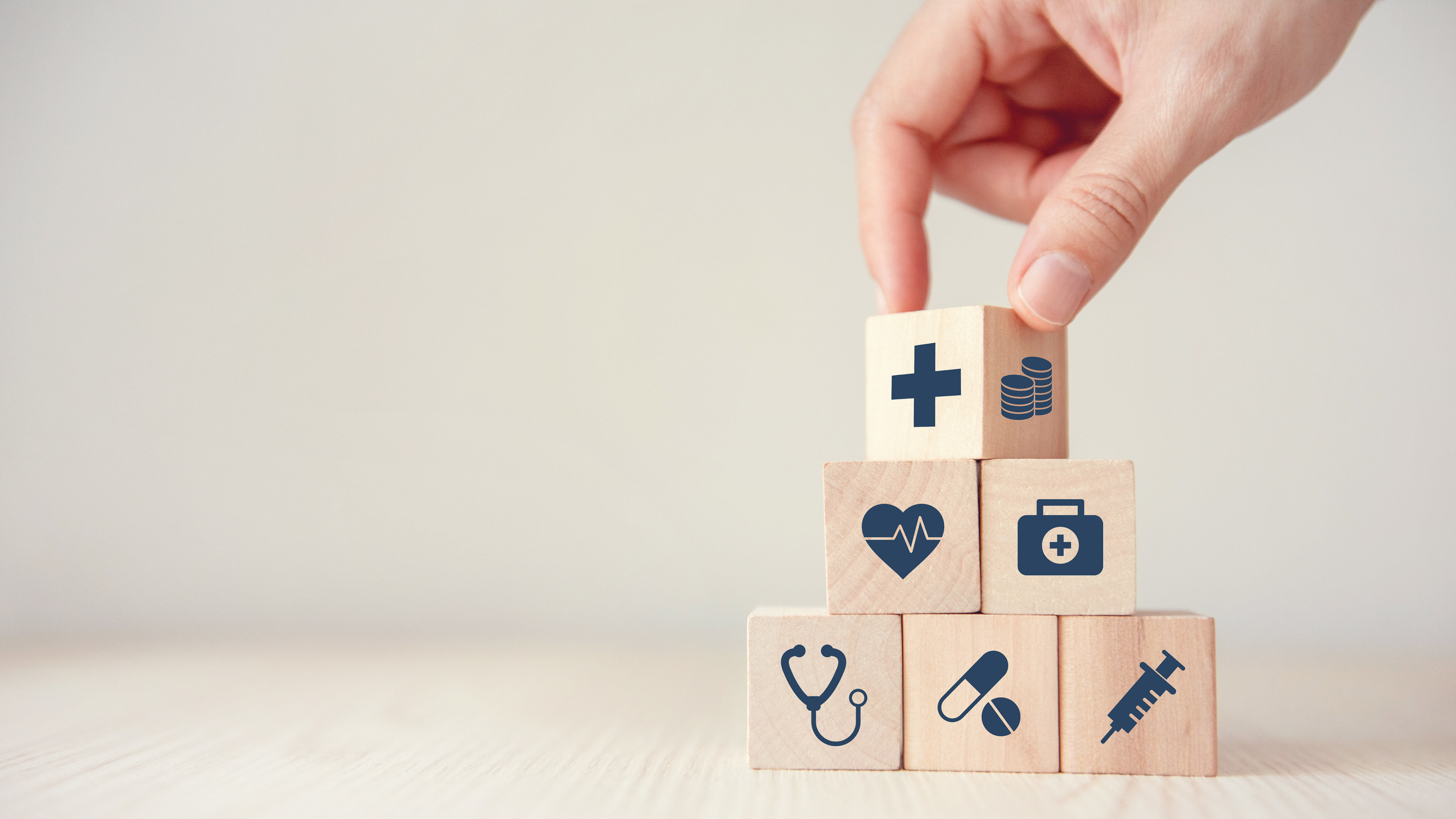 Eine Hand stapelt Holzwürfel mit Symbolen aus dem Gesundheitswesen zu einer Pyramide. Die Würfel zeigen ein Kreuz, Münzen, ein Herz mit Herzschlaglinie, einen Verbandskasten, ein Stethoskop, Medikamente und eine Spritze. Die Darstellung symbolisiert den Aufbau und die Finanzierung von Gesundheitsversorgung, medizinischen Leistungen und Arzneimittelversorgung.