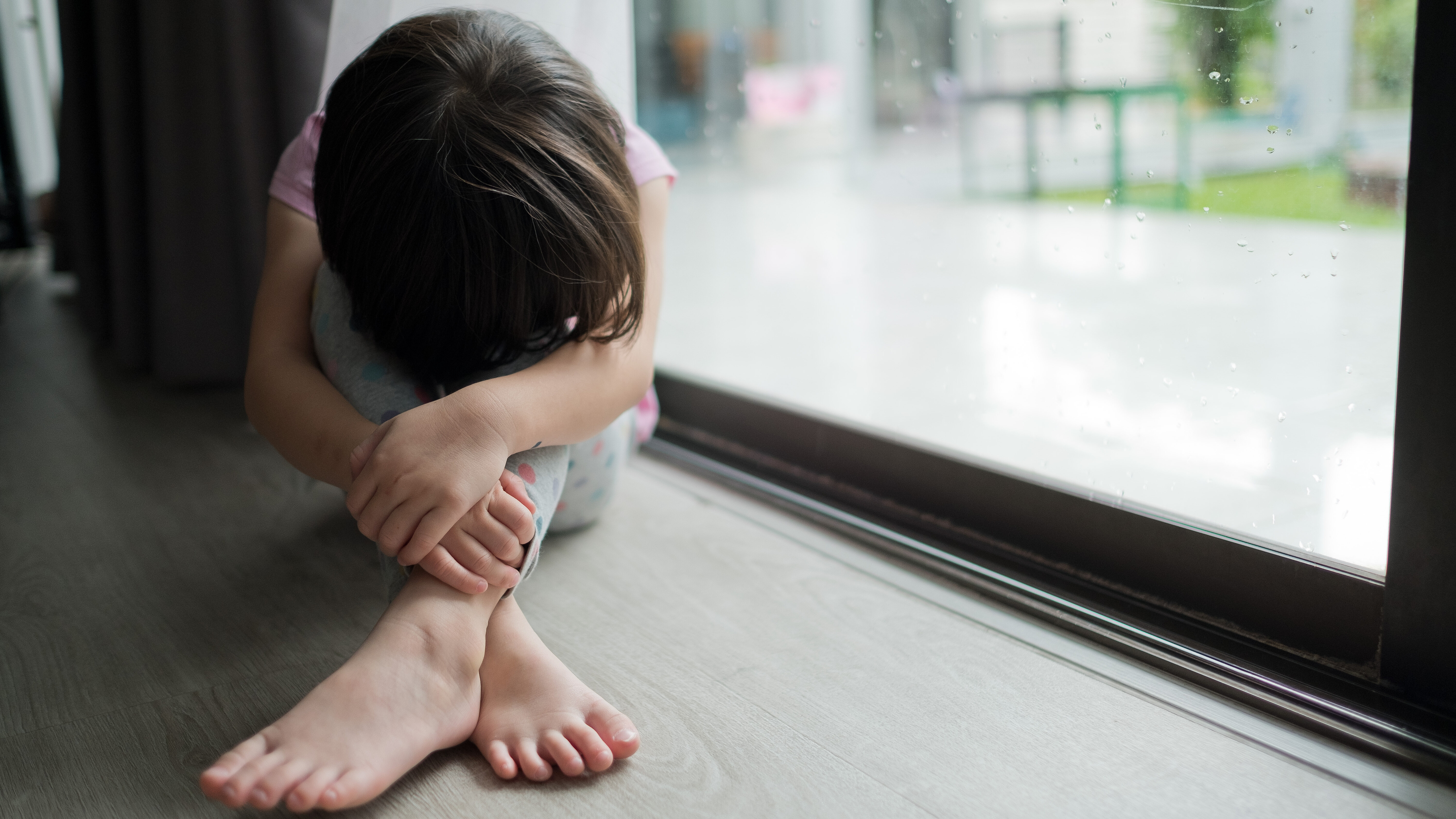 Child sitting on the floor by a window with knees drawn up