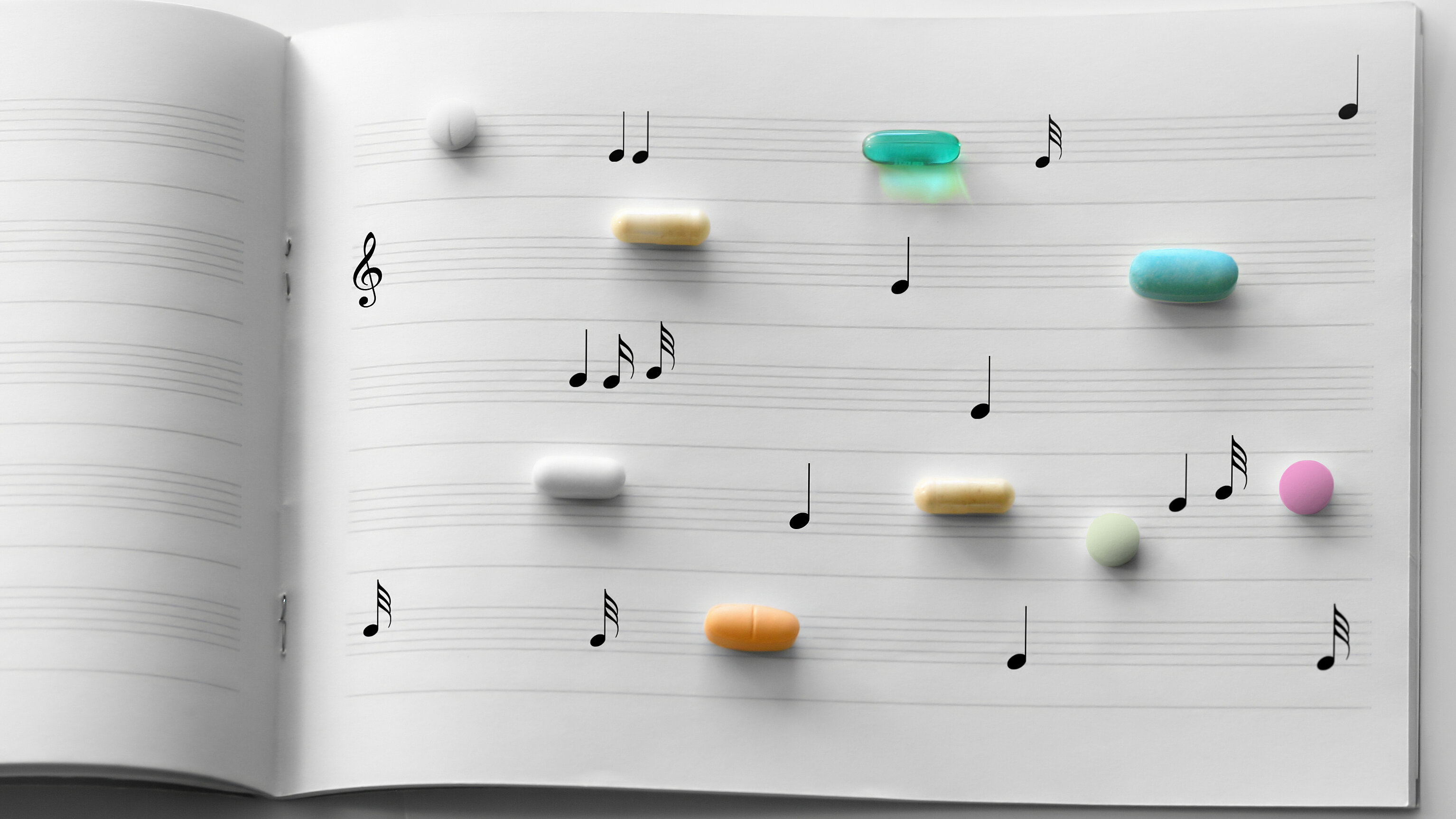 Geöffnete Notenseiten mit aufgedruckten Musiknoten, auf denen verschiedenfarbige Tabletten und Kapseln angeordnet sind. Die künstlerische Darstellung symbolisiert den Zusammenhang zwischen Musik und Gesundheit, insbesondere den Einsatz von Musiktherapie oder Musik zur Unterstützung von Behandlung und Wohlbefinden.